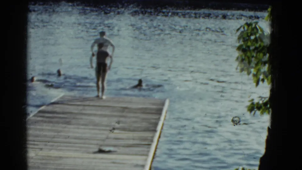 A man in swim trunks moves toward the end of a wooden pier while several swimmers remain in the lake beyond him.