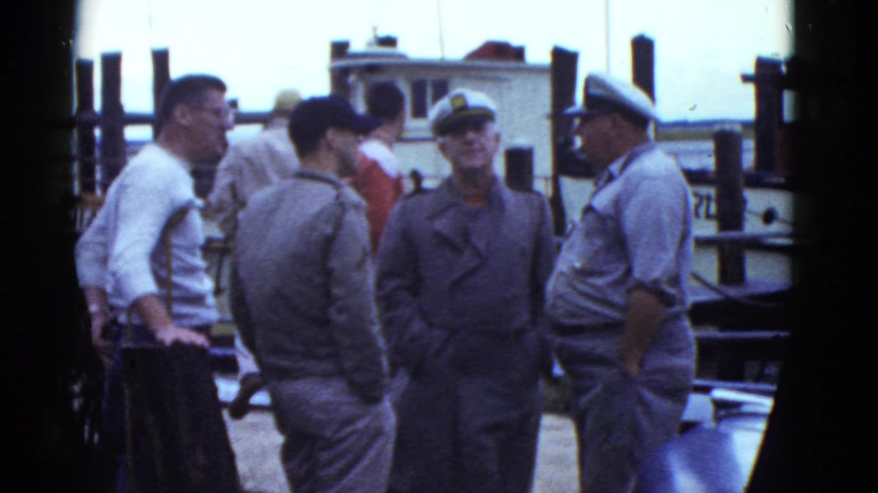 Group of men converse on a dock with a boat behind them.