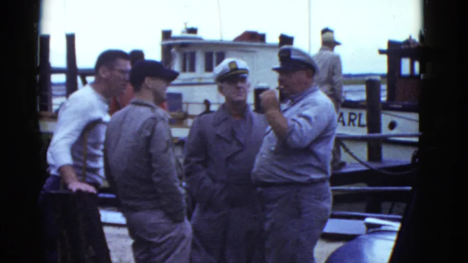 Adult men on a Boston dock continue their conversation near a small boat, with one man gesturing by his face.