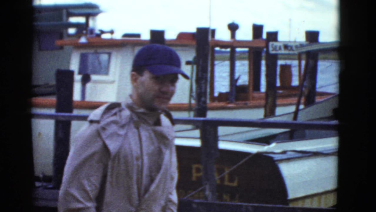 A man in a blue cap passes through a 1951 Boston dock view, with moored boats, pilings, and painted boat names behind him.