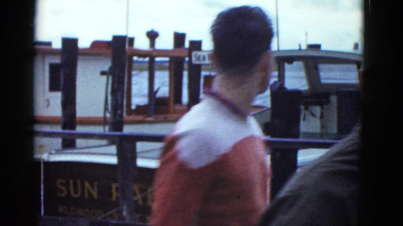 Man in a red-and-white jacket walks past a moored boat with partial lettering visible.