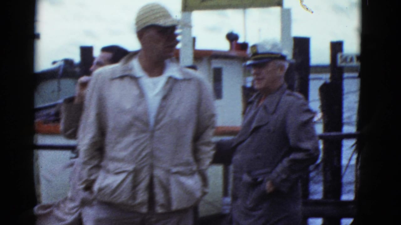 Men in caps and light jackets stand on a dock near water and posts.
