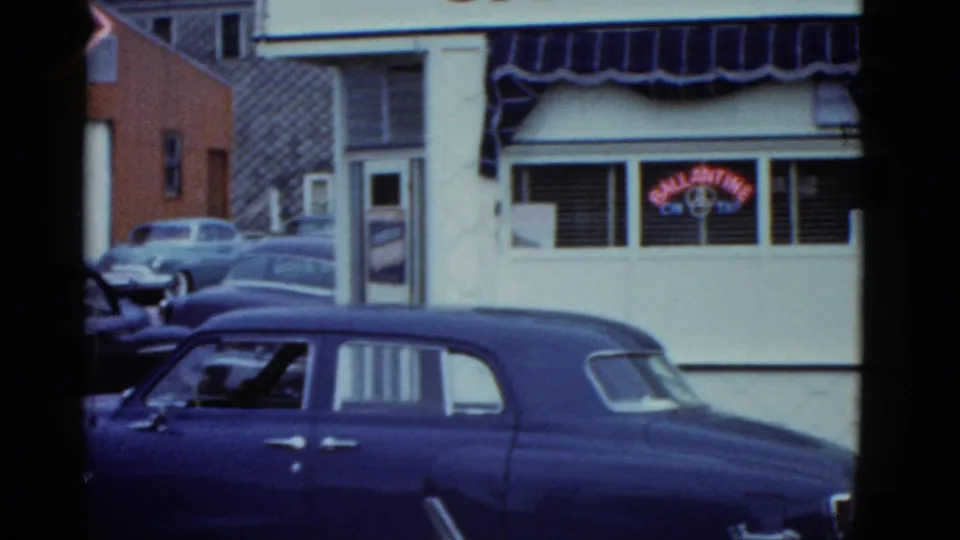 1951 Boston cafe exterior with striped awning, Ballantine neon in the window, nearby buildings, and a dark car in the foreground.