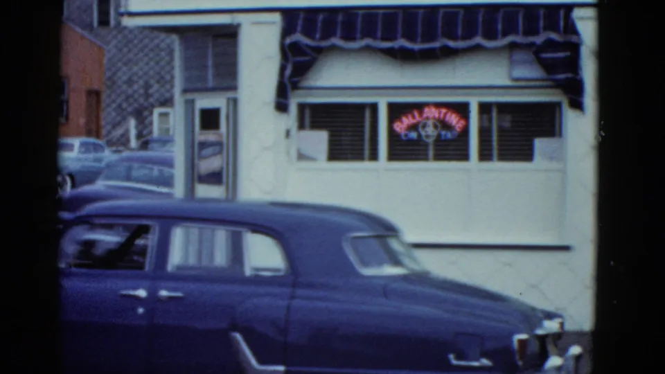 1951 Boston 8mm frame showing a blue sedan at the curb before a white storefront with awnings and a red neon beer sign.