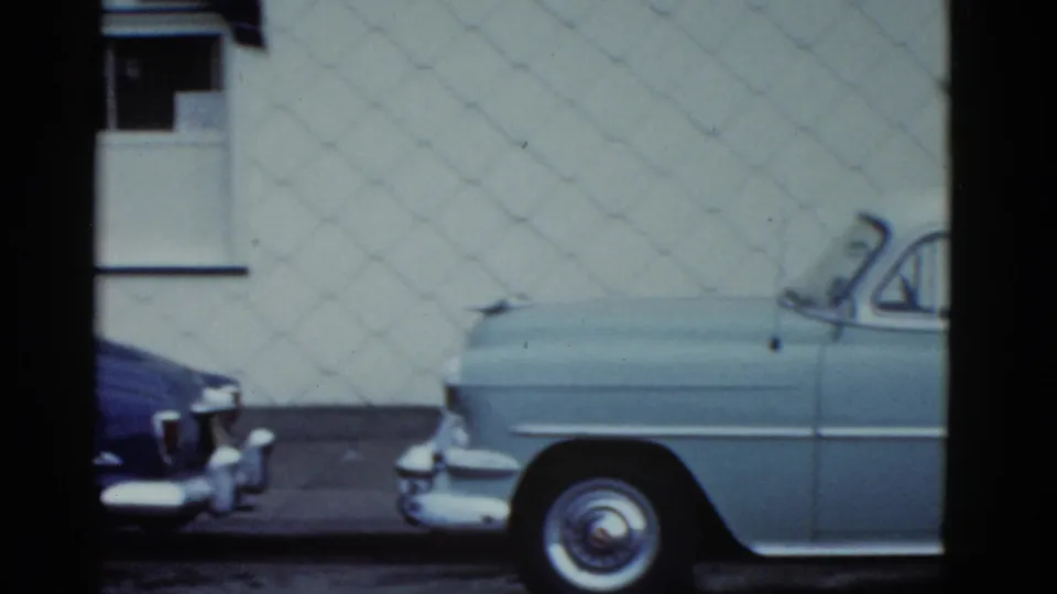 Final 1951 Boston frame showing a pale sedan at the curb, the nose of a darker car, a white wall, sidewalk, and 8mm frame borders.