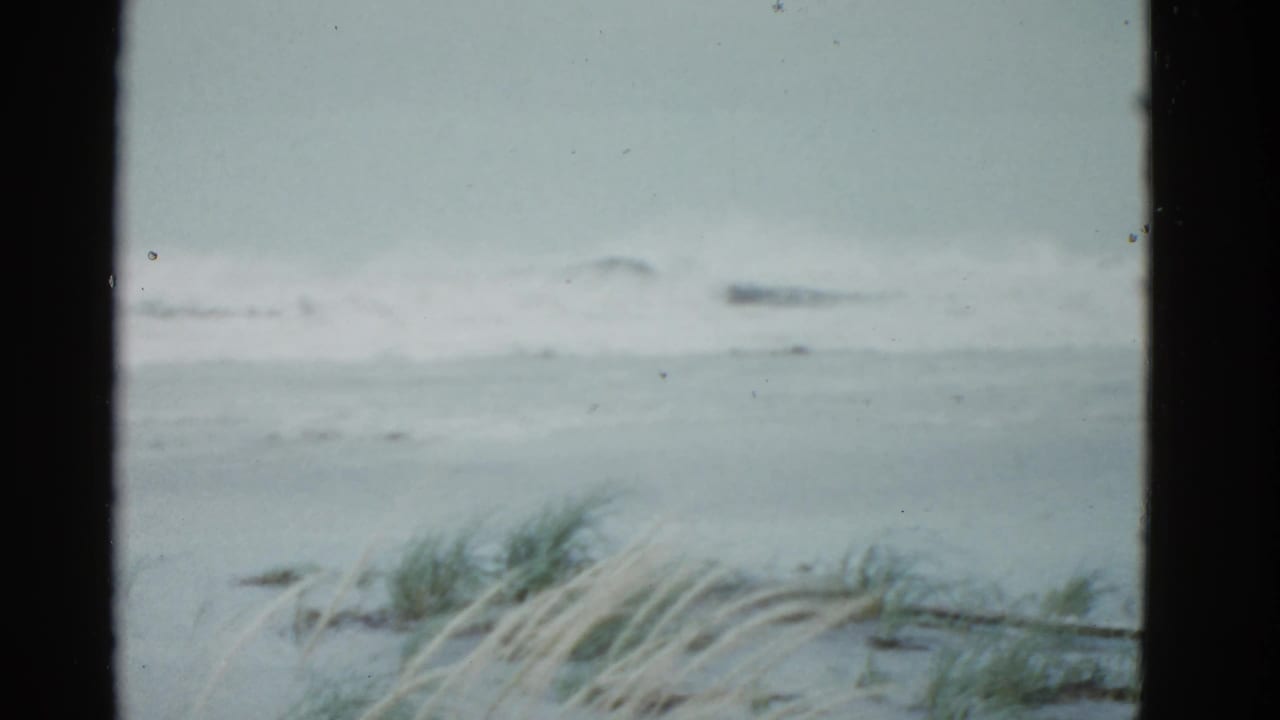 1951 Boston-area 8mm frame showing gray surf, overcast sky, and wind-blown beach grass viewed through a dark window or film-frame edge.