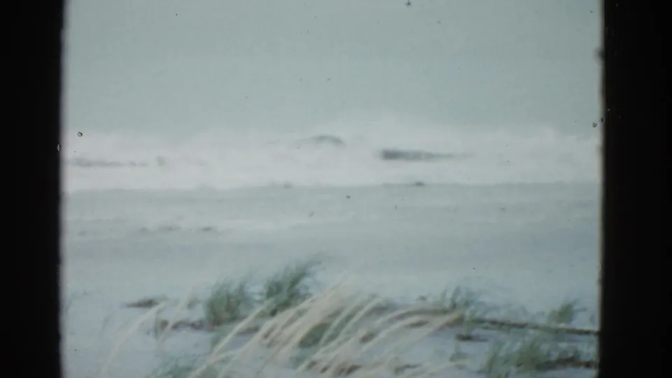 1951 Boston-area 8mm frame showing gray surf, overcast sky, and wind-blown beach grass viewed through a dark window or film-frame edge.