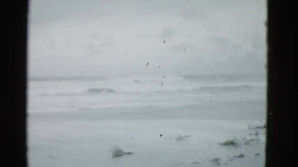 Faded 8mm frame of the Boston-area shoreline, with open water, low surf, and subdued overcast light.