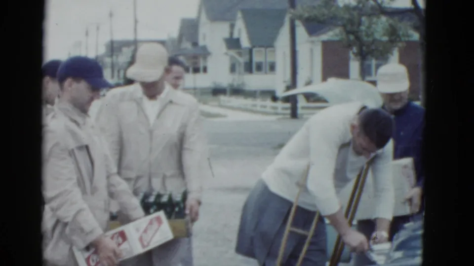 On a residential street, young men gather around a parked car while one with crutches leans toward bottles or supplies.