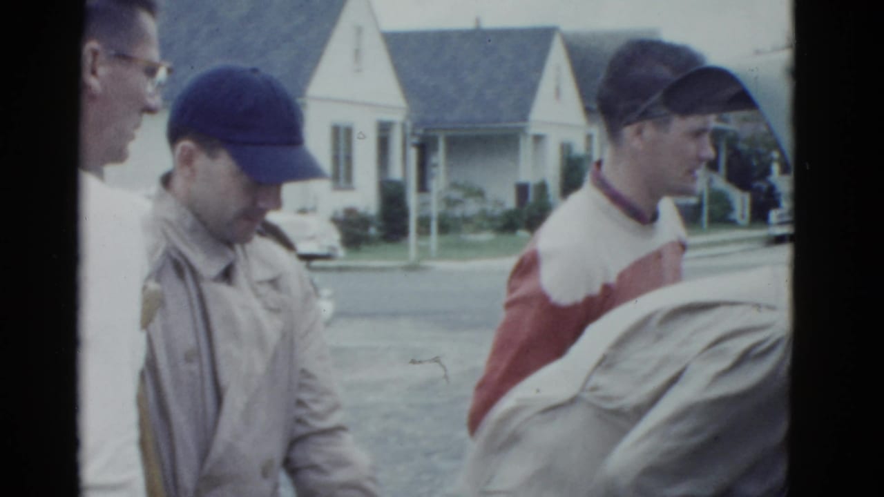 Close view of men beside a car, including a man in a red-and-white sweater.