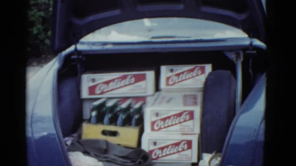 1951 Boston home-movie frame showing an open car trunk loaded with Ortlieb's beer boxes, a wooden bottle crate, glass bottles, cloth, and a spare tire.