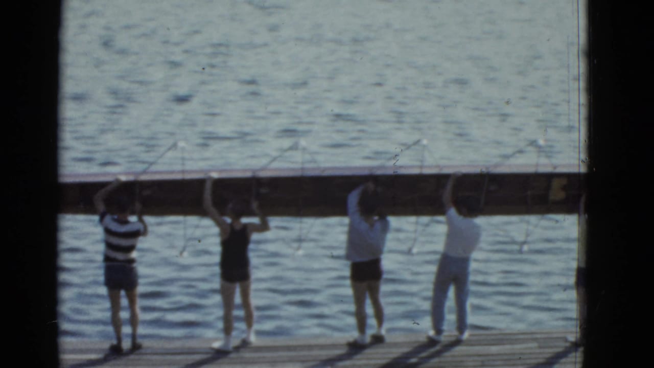 In 1951 Boston footage, several men raise a long rowing shell above their heads on a dock beside calm water.