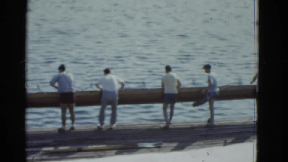 Men in athletic clothing line up beside a long rowing shell on a Boston dock, preparing to move it toward the water.