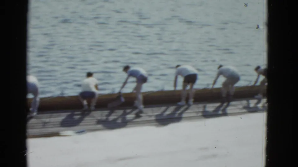 The group bends over the rowing shell at the dock edge, appearing to lower or adjust it on the water in 1951 Boston footage.