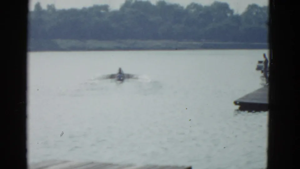 1951 Boston-area frame of a lone rower on rippled river water beside a dock edge, with trees across the far bank.