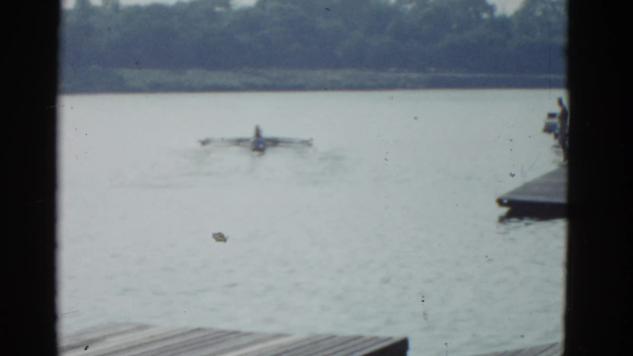 1951 Boston-area 8mm home-movie frame showing a distant rower or small craft on a lake, with dock boards at lower left and dark structural edges framing the view.