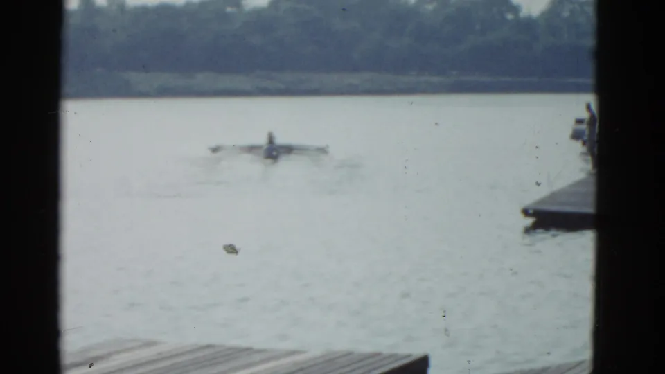 1951 Boston-area 8mm home-movie frame showing a distant rower or small craft on a lake, with dock boards at lower left and dark structural edges framing the view.