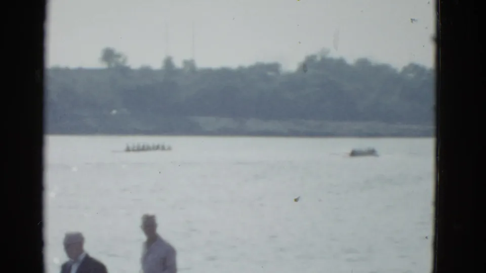 1951 Boston-area home-movie frame with two indistinct foreground people watching a lake where a rowing shell and small boat move near the far shore.