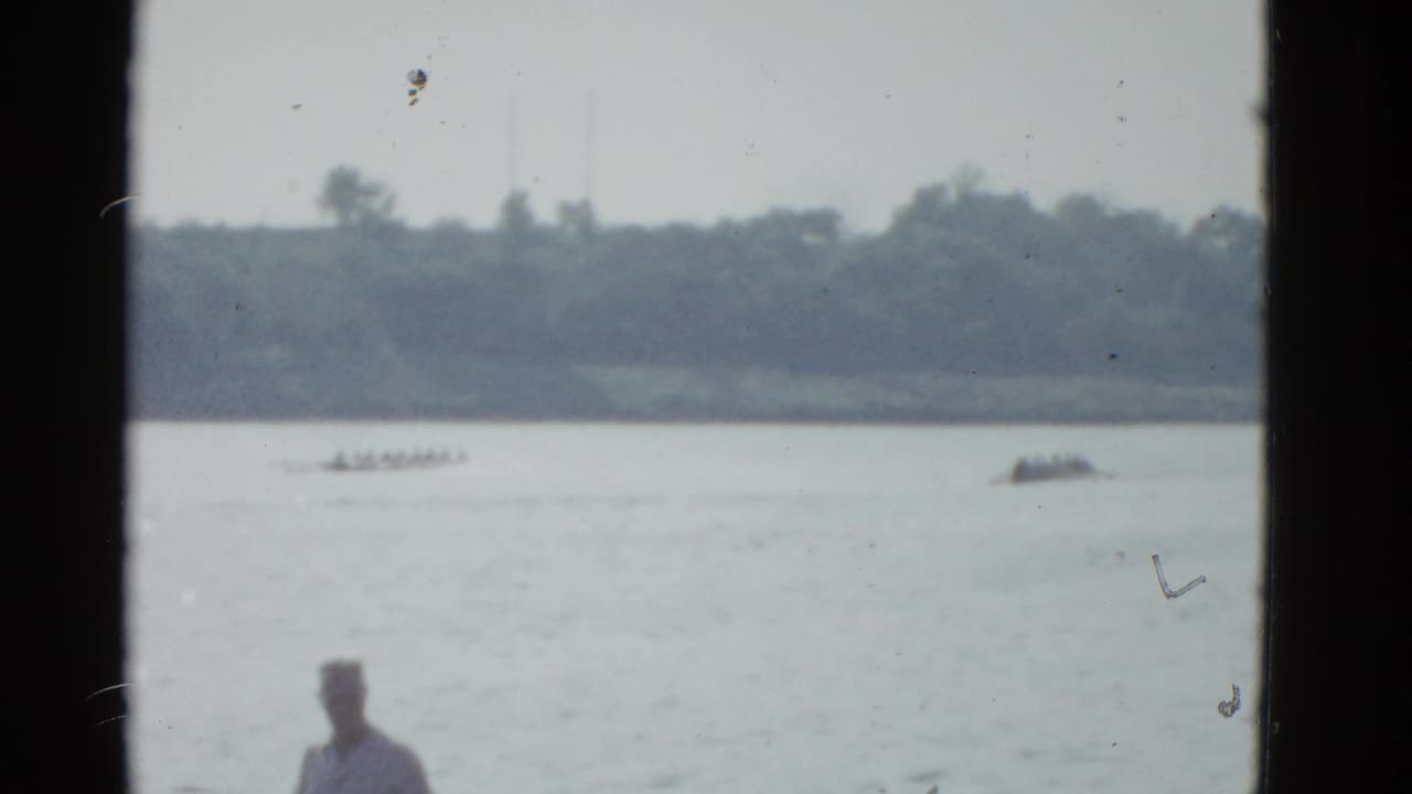 1951 Boston 8mm frame showing a distant rowing shell crossing calm water as a foreground spectator watches from the bank.