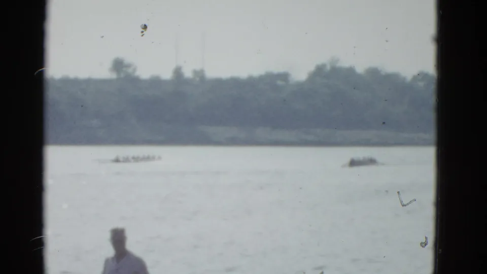 1951 Boston 8mm frame showing a distant rowing shell crossing calm water as a foreground spectator watches from the bank.