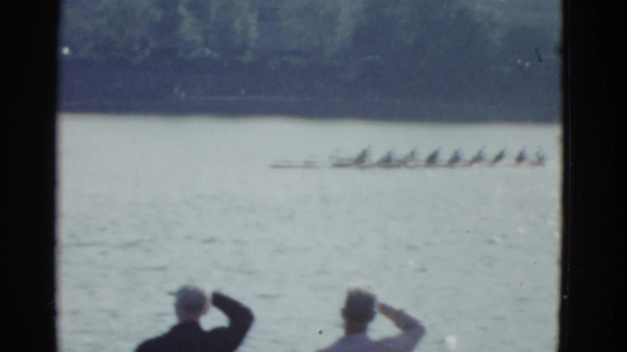 Rowing shell passes across the river as two spectators watch from the foreground.