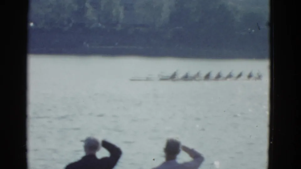 1951 Boston 8mm frame with a racing shell and synchronized rowers on the river, viewed past two foreground spectators.