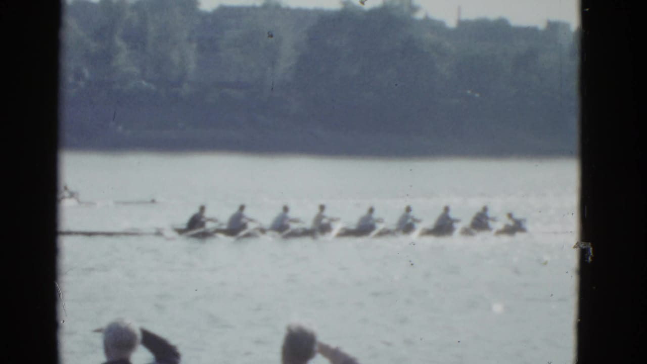 Rowing shell moves across a river with spectators partly visible in the foreground.