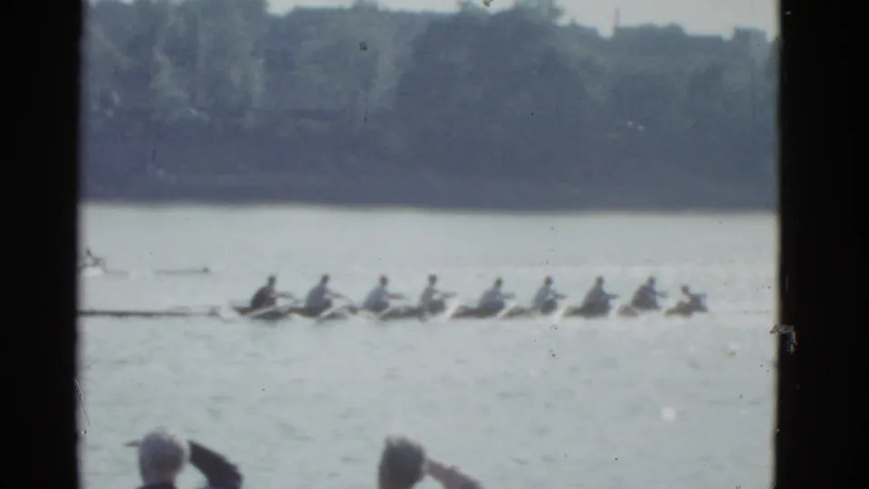 A rowing crew on a Boston river in 1951 appears mid-race, seen from a shoreline spectator viewpoint on 8mm film.