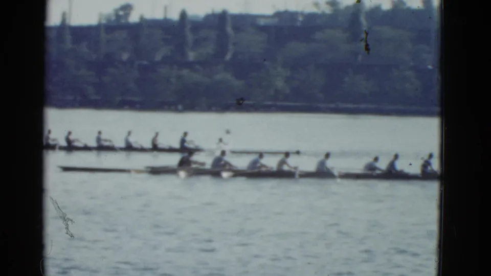 Crew boats continue along the river in Boston, with rowers and oars visible through soft, grainy 8mm home-movie footage.