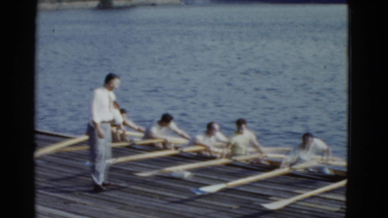Rowers in white shirts sit in a shell beside a wooden dock as a standing man supervises.