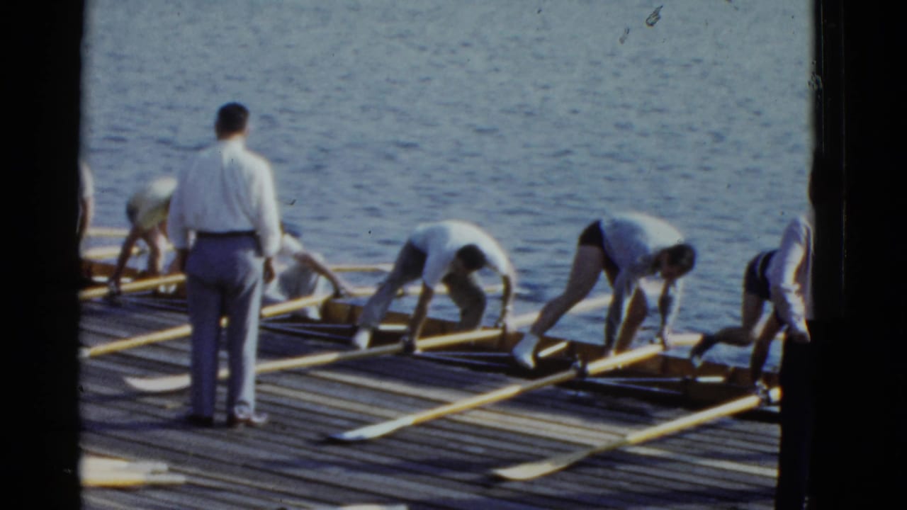 1951 Boston 8mm frame of men arranging long oars and rowing shells on a wooden pier beside the water.