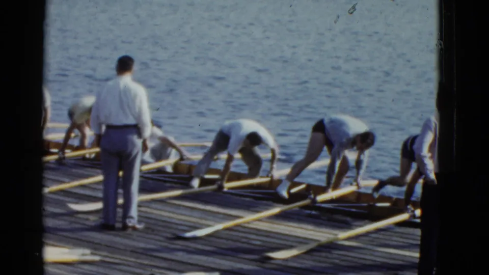 1951 Boston 8mm frame of men arranging long oars and rowing shells on a wooden pier beside the water.