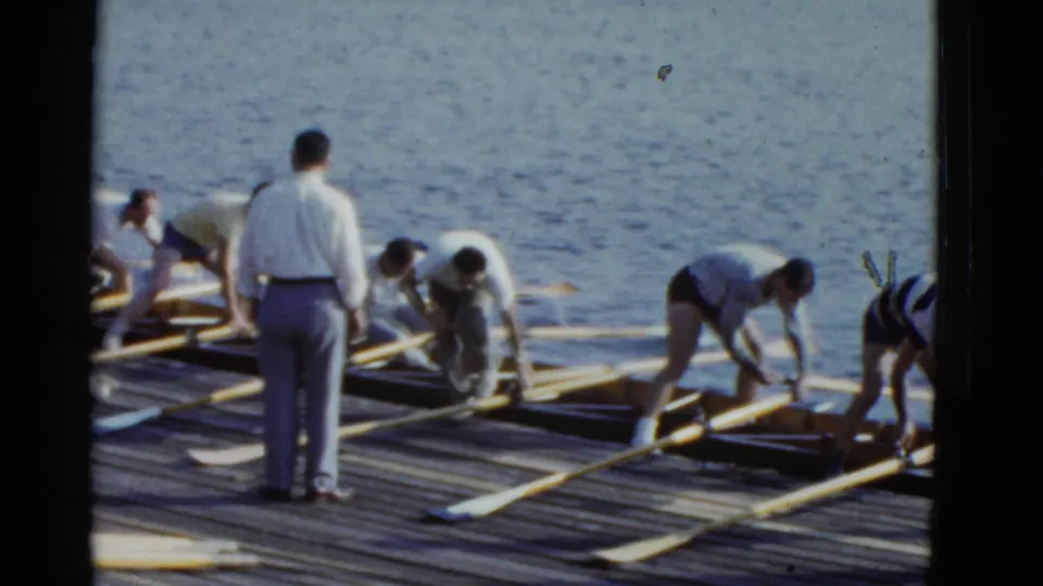 Men on a Boston pier in 1951 adjust rowing gear, with long oars laid across wooden planks and water behind them.