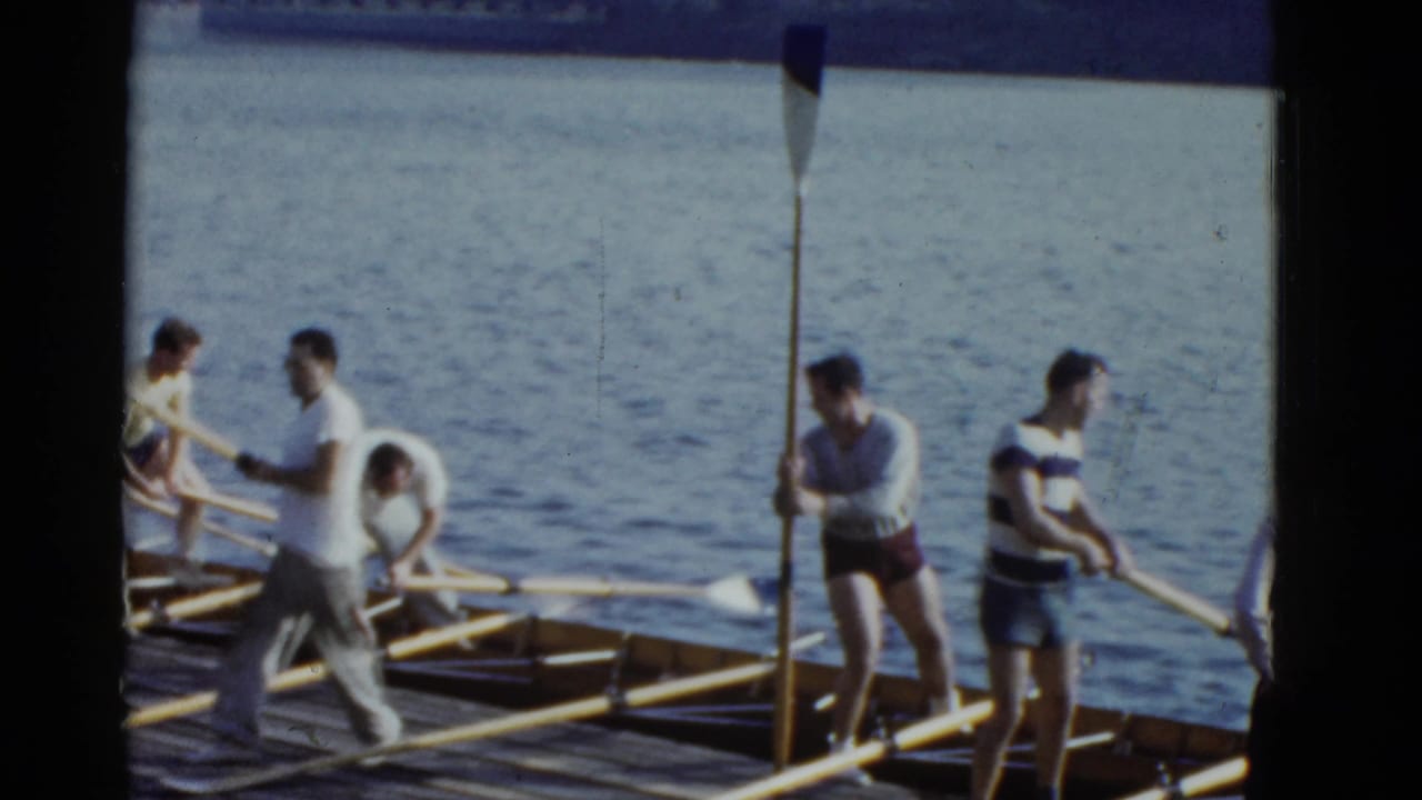 In 1951 Boston footage, men in casual clothes and athletic wear prepare oars beside a wooden rowing shell at the water's edge.