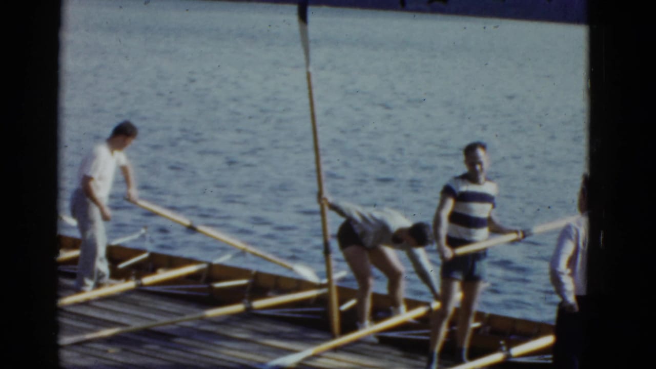 Men stand and bend over long oars on a wooden dock beside rippled water.