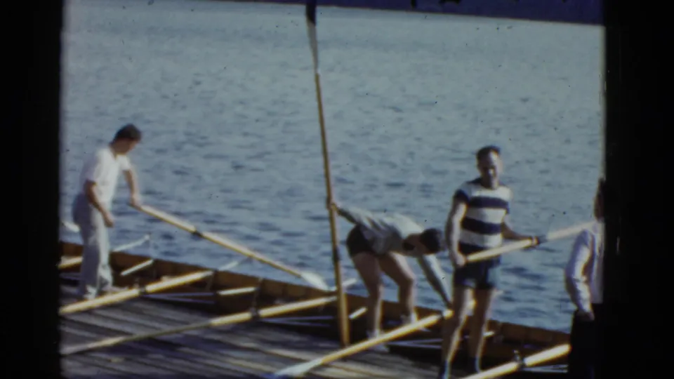 Men prepare rowing equipment on a Boston waterfront dock, with long oars laid across a wooden shell and water behind them.