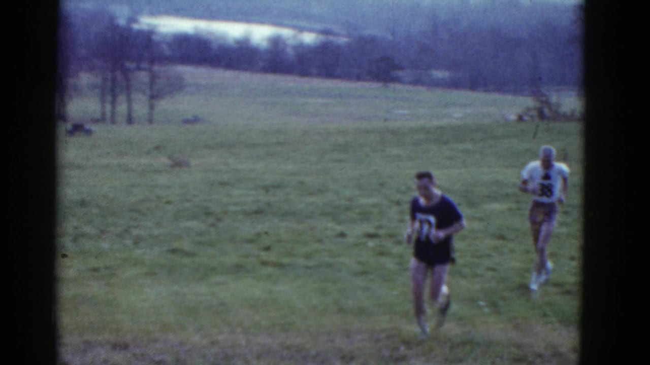 Two male runners in numbered shirts cross an open grassy field in 1951 Boston-area home-movie footage.