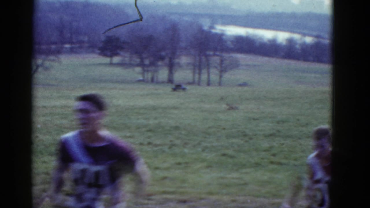 1951 Boston-area 8mm frame showing a young man in a dark athletic jersey running across an open grassy field as another figure stands nearby.