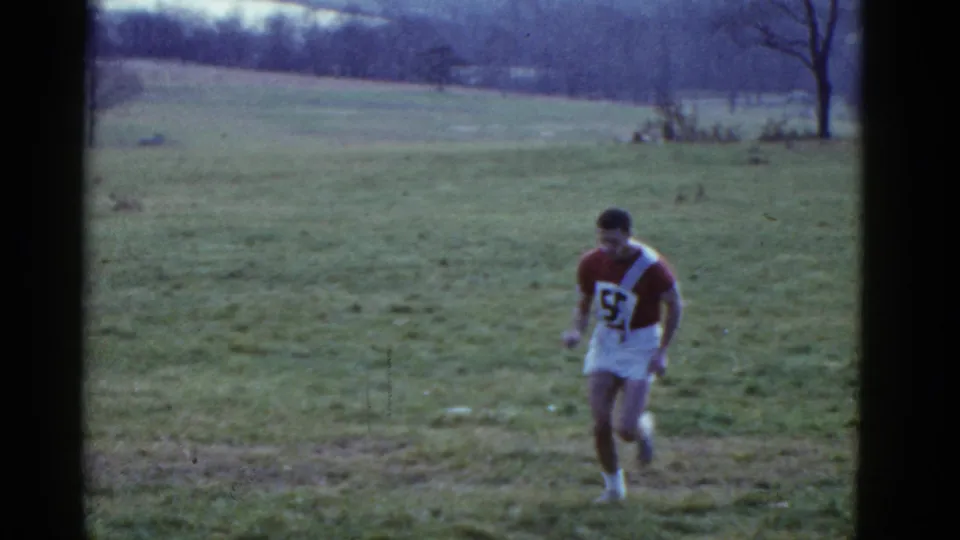 1951 Boston-area 8mm frame showing a young man in a maroon and white number 55 athletic jersey running across a grassy field near leafless trees.