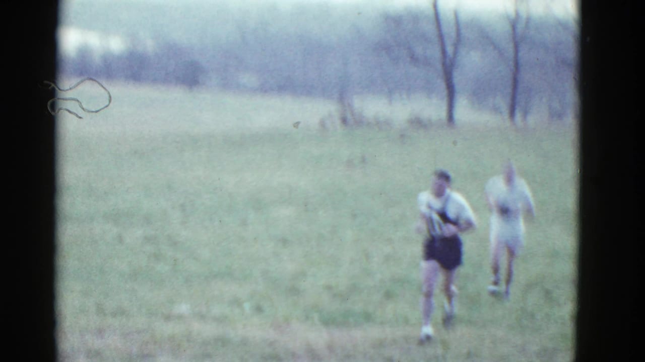 In 1951 Boston-area 8mm footage, two cross-country runners move toward the camera across an open grass field with bare trees behind them.