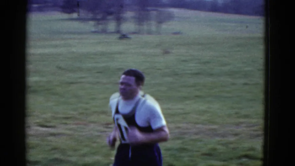 The camera catches a closer side view of a runner in a dark singlet and white shirt as he passes on the grass course.