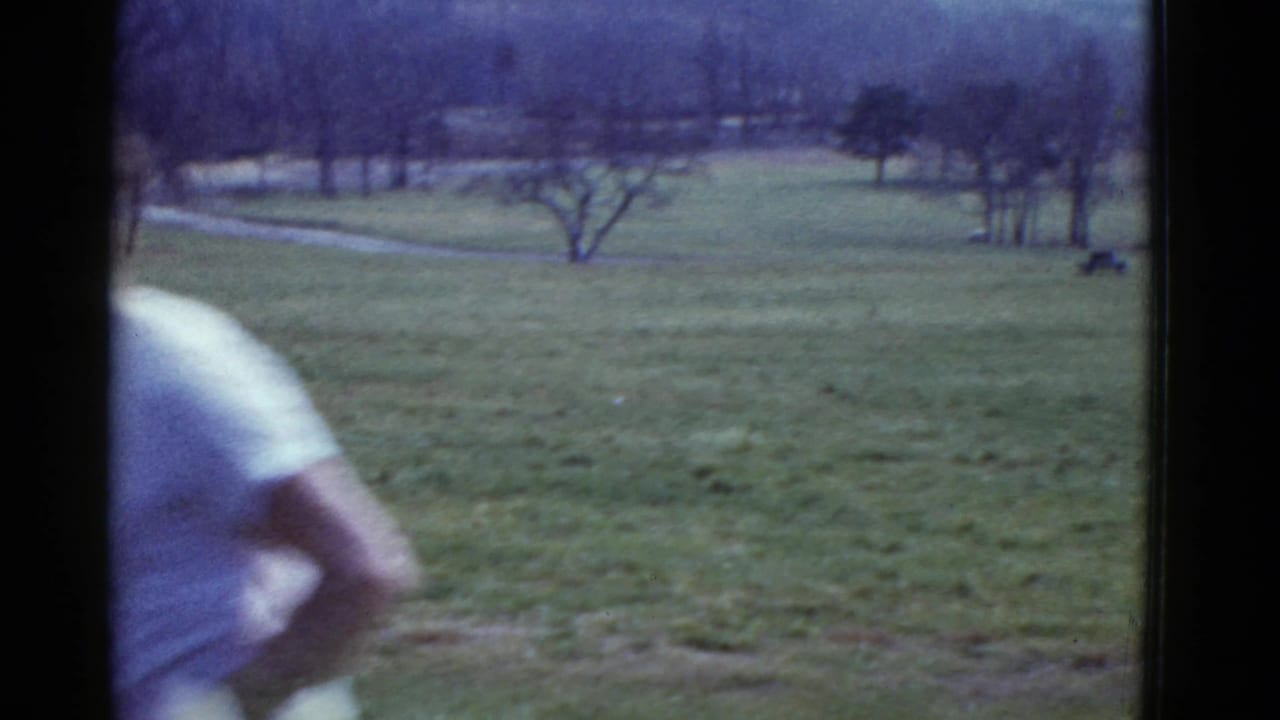 Runner partly cropped at left beside an open grassy field with bare trees in the distance.