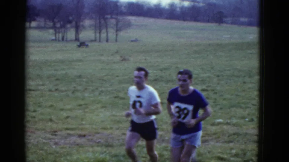 Boston 1951 8mm frame of two adult cross-country runners with visible numbers moving past the camera on a sloped lawn.