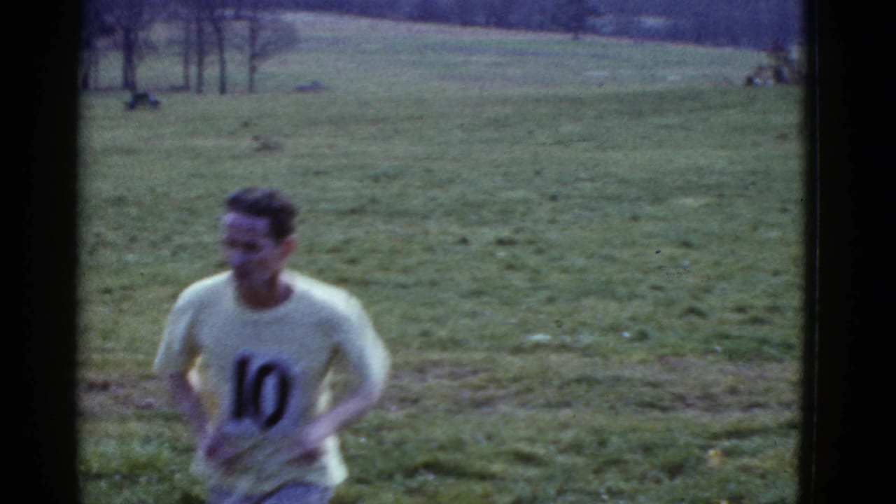 In 1951 Boston home-movie footage, a young man wearing a light numbered shirt runs near the camera across a wide grassy field.