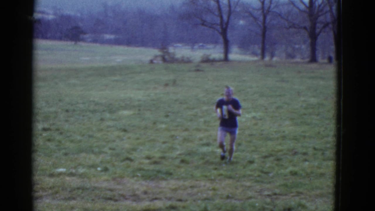 Second runner in a dark shirt approaches across a wide grassy field.