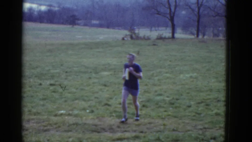 The dark-shirted runner continues toward the camera across the open field, framed by 8mm film edges and distant bare trees.