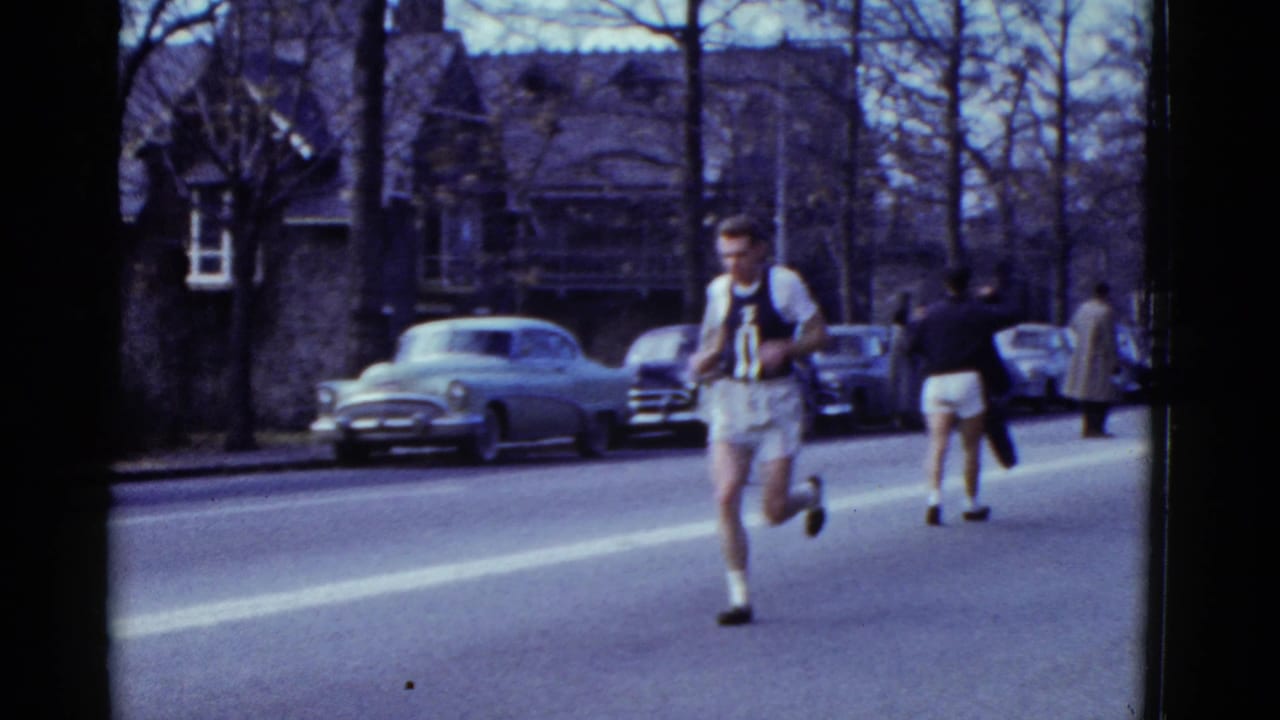 Male runner in a dark singlet and white shorts runs along a residential street with parked cars behind him.
