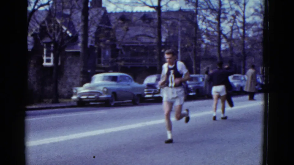 1951 Boston home-movie frame showing a male road runner passing parked mid-century cars on a tree-lined residential street.