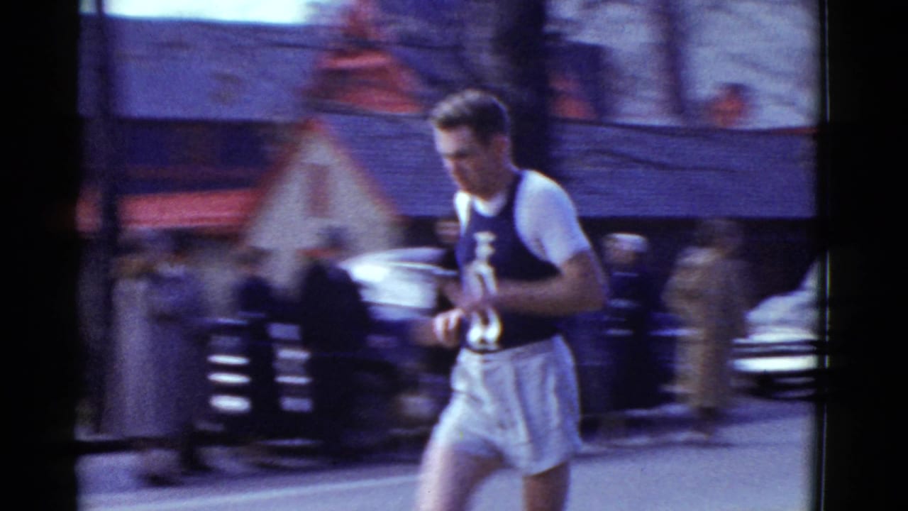 Runner in a dark-and-white uniform passes close to the camera with blurred spectators and cars behind him.