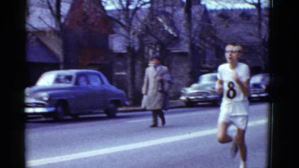 Male runner with bib number 8 moves through a Boston residential street race, with a parked car and bundled pedestrian nearby.